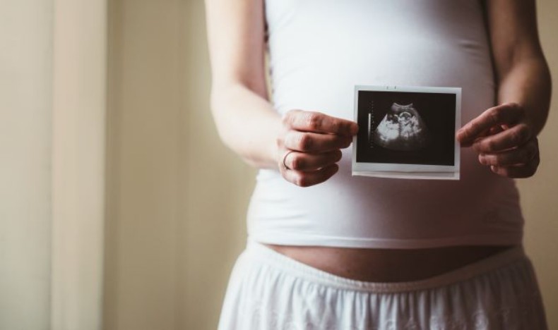 A pregnant person holds up a printed ultrasound image over her belly.