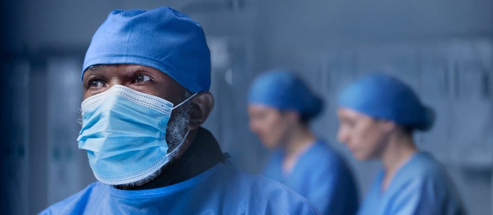 A male healthcare professional wearing a mask is preparing for a medical procedure in an operating room. Two more clinicians are in the background.