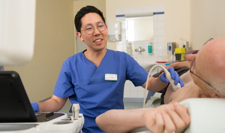 A medical professional in blue scrubs performs an ultrasound examination on a patient lying on a medical bed. The clinician holds an ultrasound probe against the patient&rsquo;s chest while looking toward a monitor. The patient&rsquo;s arm is visible in the foreground. Medical equipment and tiled walls in the background indicate a clinical examination room.