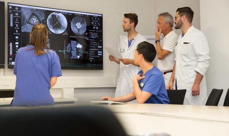 A group of healthcare professionals gathers in a meeting or consultation room, focusing on a large wall-mounted medical display showing detailed imaging scans. One person in blue scrubs stands closest to the screen with their back to the camera, while others in white coats and blue scrubs review documents and discuss the images collaboratively.