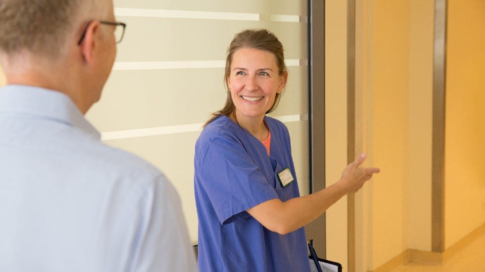 A healthcare worker in blue scrubs stands in a hospital corridor, smiling and gesturing with an open hand as if giving directions or welcoming someone. A person in a light-colored shirt is seen from behind in the foreground, facing her. The setting appears clean and modern, with glass walls and neutral tones.